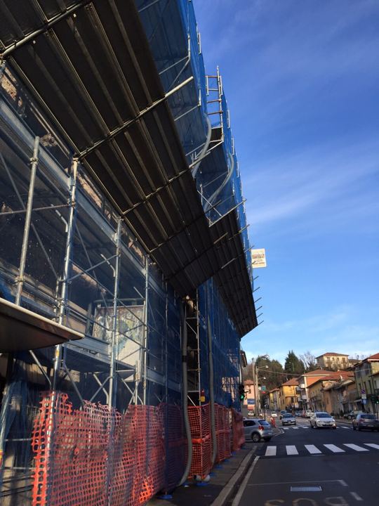 Ponteggio con rete blu e delimitazione di cantiere su viabilità pubblica.